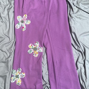 Może przedstawiać: Lawendowe spodnie dresowe z trzema dużymi kwiatowymi aplikacjami. Każdy kwiat wykonany jest z kolorowej tkaniny kwiatowej z szarym środkiem. Spodnie mają elastyczny pas i luźny krój.