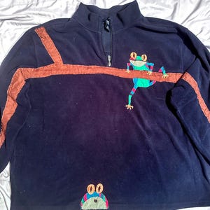 May include: Navy blue fleece pullover with a high collar and a quarter-zip closure. The pullover features a brown stripe design and a colorful frog applique. A second frog applique is at the bottom of the shirt.
