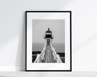May include: Black and white framed print of a lighthouse at the end of a wooden pier. The lighthouse has a black cupola and white walls. The pier extends into the ocean, with a cloudy sky in the background.