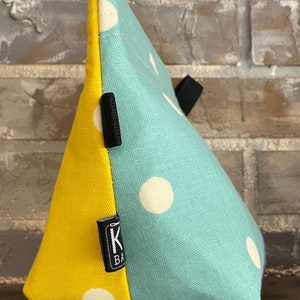 May include: A yellow and teal fabric pouch with white polka dots. The pouch has a black zipper and a black label with the letter "K" and the word "Bags" on it.