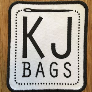 May include: White square patch with black border and stitching. The patch has a black needle and thread design at the top, the letters "KJ" in large black font, and the word "BAGS" in smaller black font.