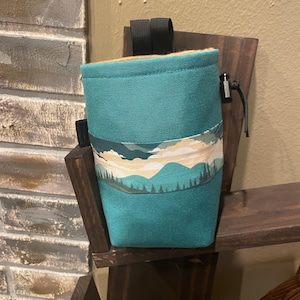 Può includere: Borsa per magnesite da arrampicata color verde acqua con un design a paesaggio montano. La borsa ha una chiusura con coulisse e una cinghia nera.