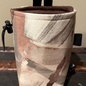 May include: A brown and beige patterned drawstring bag with a brown fleece lining. The bag has two black straps at the top and a black drawstring closure.