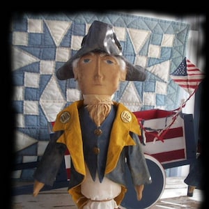 May include: A handmade rag doll dressed as a colonial soldier. The doll is wearing a blue jacket with yellow lapels, a white shirt, and a black tricorne hat. The doll is standing in front of a red, white, and blue quilt.