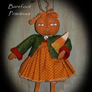 May include: A handmade doll with a pumpkin-shaped head, blue eyes, and a green jacket with red trim. The doll wears an orange dress with a white floral pattern and holds a candy corn. The text "Barefoot Primitives" is in the upper left corner.