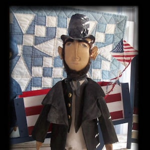 Puede incluir: Una muñeca de trapo hecha a mano vestida como Abraham Lincoln. La muñeca lleva un abrigo negro, una camisa blanca y un sombrero de copa negro. La muñeca está de pie frente a una colcha roja, blanca y azul y un carro de madera.