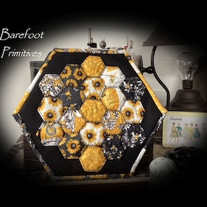 May include: A handmade, hexagonal quilted item with a black border. The center features yellow sunflower and black floral patterned fabric. The text "Barefoot Primitives" is in the upper left corner. A vintage lamp and other decorative items are in the background.