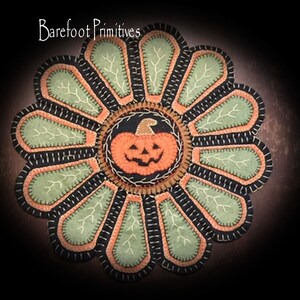Può includere: Un tappeto in feltro nero, verde e arancione con un disegno di zucca al centro. Il tappeto ha la forma di un fiore con 12 petali. Il testo "Barefoot Primitives" è in cima al tappeto.