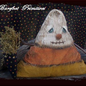 May include: A handcrafted Halloween decoration shaped like a candy corn. The decoration has a white top, orange middle, and yellow bottom. It features a stitched mouth, button eyes, and a twig arm. The background is a black fabric with colorful polka dots.
