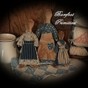 May include: Three handmade rag dolls dressed in vintage-style clothing. The dolls are standing on a blue and white patchwork quilt. The dolls are wearing blue and white striped skirts, a brown skirt with blue and white polka dots, and a blue and white striped dress. The dolls are all wearing white bonnets. The text "Barefoot Primitives" is visible in the background.