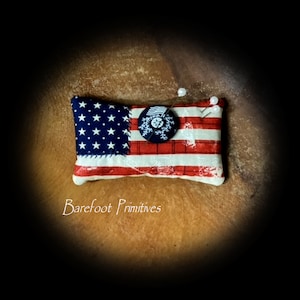 May include: A small, red, white, and blue fabric pincushion in the shape of the American flag. The pincushion has a blue button with a white snowflake design in the center. The pincushion is sitting on a brown surface.