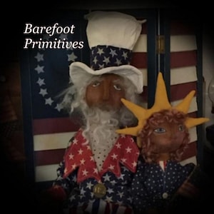 May include: Two handmade patriotic dolls, one resembling Uncle Sam with a tall hat and star-spangled jacket, the other with a sunburst crown. The background features a painted American flag and the text "Barefoot Primitives".