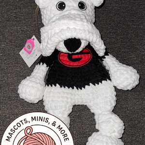 Puede incluir: Peluche de bulldog de ganchillo blanco y negro hecho a mano. El bulldog tiene un logotipo "G" negro y rojo en el pecho. El juguete tiene ojos negros grandes y una nariz negra. El producto es de "Mascots, Minis, & More".