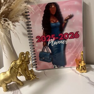 May include: A spiral-bound 2025-2026 planner with a pink cover featuring a fashion illustration of a woman in a denim dress. The planner is next to gold elephant figurines and a vase with dried pampas grass.