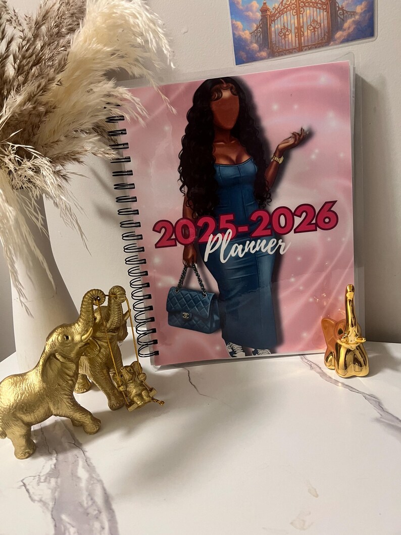 May include: A spiral-bound 2025-2026 planner with a pink cover featuring a fashion illustration of a woman in a denim dress. The planner is next to gold elephant figurines and a vase with dried pampas grass.