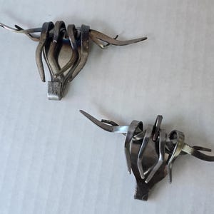 Puede incluir: Dos esculturas metálicas con forma de cráneos de toro longhorn, elaboradas con tenedores plateados doblados y soldados. Cada pieza presenta dientes curvados que se extienden hacia afuera para asemejarse a cuernos, con el mango del tenedor.