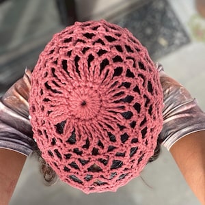 Peut inclure: Un bonnet en crochet de couleur rose poussiéreux. Le bonnet a un design en dentelle avec un motif circulaire au centre.