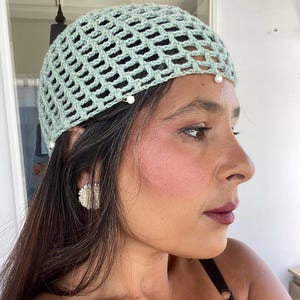 Puede incluir: Un gorro de crochet verde claro con un patrón de rejilla y detalles de perlas blancas.