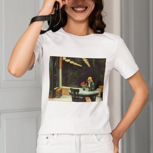 May include: White t-shirt with a graphic print of a painting depicting a woman sitting at a table in a diner. The painting is in a dark blue, green, and yellow color scheme.