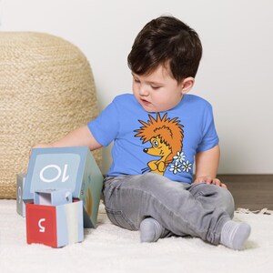 Könnte beinhalten: Ein kleines Kind trägt ein hellblaues T-Shirt mit einem Comic-Igel, der Blumen hält, das auf der Vorderseite aufgedruckt ist. Das Kind sitzt auf einem hellblauen Teppich und spielt mit bunten Blöcken.