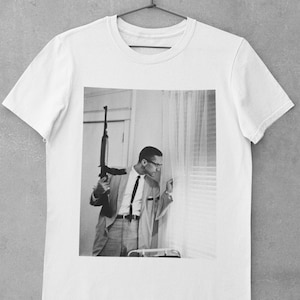 Puede incluir: Una camiseta blanca con una foto en blanco y negro de un hombre sosteniendo un rifle y mirando por una ventana. El hombre lleva un traje y una corbata.