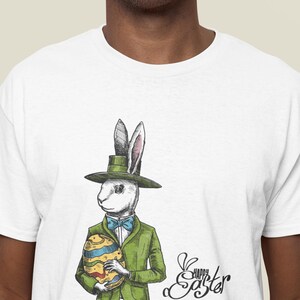 Puede incluir: Camiseta blanca con una ilustración de dibujos animados de un conejo con un sombrero verde y sosteniendo un huevo de Pascua decorado. El texto "Feliz Pascua" está escrito en cursiva debajo del conejo.