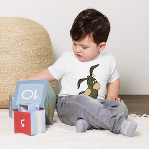 May include: A young child wearing a white t-shirt with a green cartoon donkey on it. The child is sitting on a white surface and playing with colorful blocks.