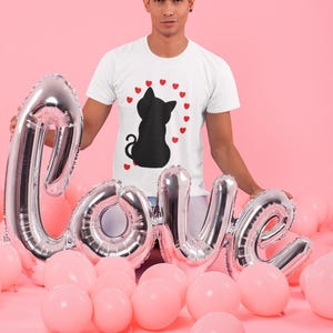 May include: A white t-shirt with a black silhouette of a cat surrounded by red hearts. The person wearing the shirt is holding a silver balloon that says "Love" in large letters. The background is pink.