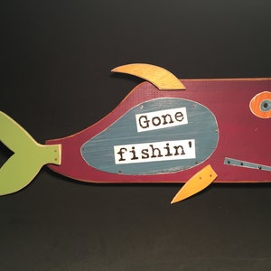 May include: A wooden fish sculpture painted in shades of red, green, yellow, and blue. The fish has a large eye and a sign that reads "Gone fishin'" in black lettering.