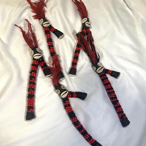 Puede incluir: Cuatro objetos rituales hechos a mano. Cada uno presenta un eje central de cuentas en negro y rojo alternados, con un mango en forma de T. La parte superior tiene una concha de cauri y plumas rojas. La base es de forma negra.