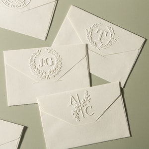 Puede incluir: Cuatro sobres blancos con diferentes monogramas en relieve. Los monogramas son "JG", "T", "AC" y un diseño floral.