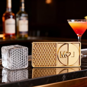 Puede incluir: Un molde para cubitos de hielo dorado con un patrón de panal y las iniciales "M♥J" junto a un cubito de hielo transparente con un patrón similar. Un cóctel y botellas están en el fondo.