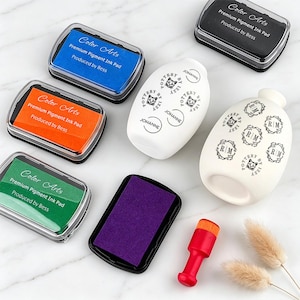 May include: A collection of five rectangular Color Arts premium pigment ink pads in blue, orange, green, black, and purple, alongside two white ceramic pottery pieces with black stamped text and a small red handled stamp tool with an orange base.