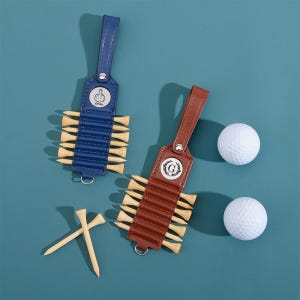 May include: Two golf tee holders with leather straps, one blue and one brown, each holding wooden tees. Two white golf balls are also present. The blue holder has a silver emblem with a hand gesture. The brown holder has a silver emblem with a letter G.