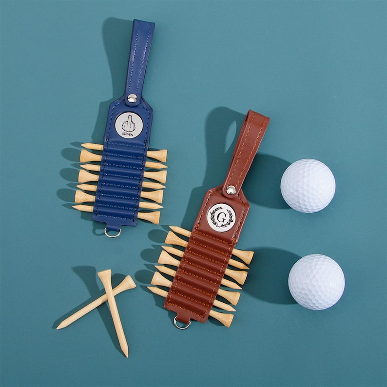 May include: Two golf tee holders with leather straps, one blue and one brown, each holding wooden tees. Two white golf balls are also present. The blue holder has a silver emblem with a hand gesture. The brown holder has a silver emblem with a letter G.