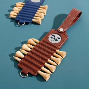Op de afbeelding: Bruin lederen golf tee-houder met de tekst "KIEVEN" en een golfclublogo. De houder bevat verschillende houten golf tees. Een blauwe lederen golf tee-houder met houten tees staat op de achtergrond.