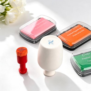 May include: A collection of art supplies featuring ink pads in pink, orange, and green, a white ceramic cup with a blue floral design, and a red stamp with an orange stamp pad. The ink pads are labelled "Color Arts" and "Premium Pigment Ink Pad."