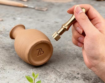Puede incluir: Un pequeño jarrón de terracota sin esmaltar con cuerpo redondeado y cuello estrecho. La base del jarrón tiene un diseño "B" estampado. Se sostiene una herramienta de latón cerca del jarrón, lo que sugiere un proceso de alfarería o artesanía. El fondo incluye herramientas de alfarería y arcilla.