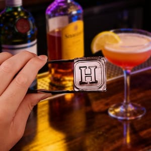May include: A silver ice stamp with the letter "H" is held by tongs. A cocktail with a lemon garnish sits on a wooden bar. Bottles of liquor are in the background.