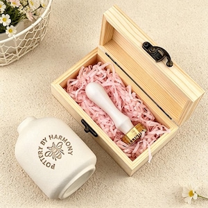May include: A wooden box with a brass and white wax seal stamp, nestled in pink paper. Beside it, a white ceramic bottle with the text "POTTERY BY HARMONY" and a bee illustration.