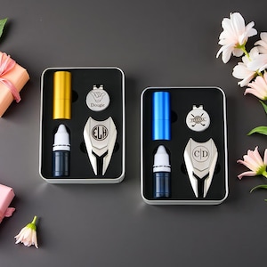 May include: Two golf gift sets in black metal boxes. Each set includes a gold or blue cylindrical item, a small bottle, a divot tool, and a ball marker. The divot tools are silver with white accents and the initials "ELD" and "CD".