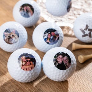 May include: Six white golf balls with personalized photos printed on them. The photos include a cat, a couple, and a group of people. The golf balls are on a wooden surface.