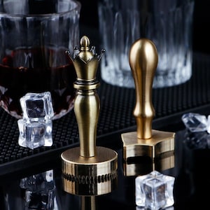 May include: Two antique brass wax seal stamps. One is shaped like a chess queen with a crown, the other has a rounded handle and a star-shaped base. Clear ice cubes and a glass of dark liquid are in the background.