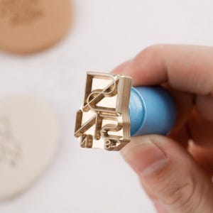 May include: A brass stamp with the letters "RUN" in a stylized font. The stamp has a light blue handle. The stamp is held in a person's hand. A light brown object is in the background.