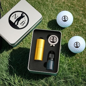 Puede incluir: Una caja de regalo de metal plateado con un diseño de golf en blanco y negro. La caja contiene una botella dorada, un marcador de bola de golf negro y plateado y una pequeña botella de líquido. Dos bolas de golf blancas con un diseño de golf en blanco y negro están sobre la hierba verde.