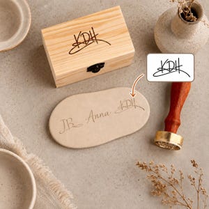 May include: A rectangular light-coloured wooden box with a black metal latch, featuring the engraved letters KDH. Beside it, an oval-shaped beige clay piece displays the engraved names JB, Anna, and KDH, alongside a wooden wax seal stamp.