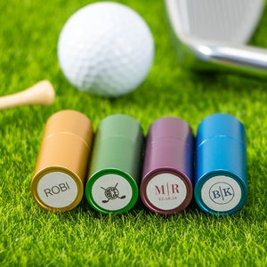 May include: Four cylindrical metal golf ball markers in gold, green, purple, and blue. Each marker features a white circular top with custom engravings: the name ROBI, a golf club icon with the text ACE, the initials M|R with 12.10.24, and the initials BK.