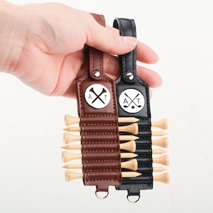 May include: Two golf tee holders, one brown and one black, each with a leather strap and a metal loop. The holders feature a circular emblem with a golf ball, crossed clubs, and the letters "A" and "T". Each holder contains several wooden golf tees.