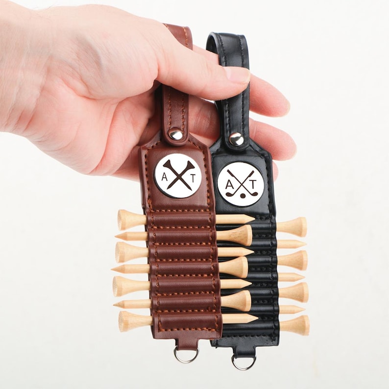 May include: Two golf tee holders, one brown and one black, each with a leather strap and a metal loop. The holders feature a circular emblem with a golf ball, crossed clubs, and the letters "A" and "T". Each holder contains several wooden golf tees.