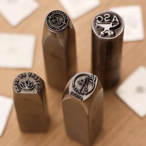 Könnte beinhalten: Vier Metallstempel mit Holzgriffen werden gezeigt. Die Stempel haben verschiedene Designs, darunter eine Lotusblume, die Buchstaben "A" und "S" mit einem Amboss und die Wörter "handmade" und "boller" in einem kreisförmigen Muster. Die Stempel befinden sich auf einer Holzoberfläche.